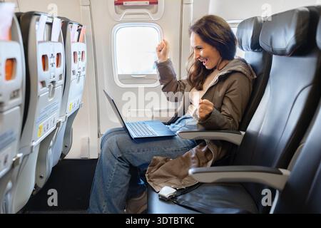 Femme caucasienne heureuse célébrant le succès tout en utilisant un ordinateur portable sur le siège de l'avion Banque D'Images