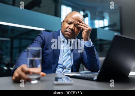 Homme d'affaires noir fatigué tenant le verre d'eau et touchant la tête dans le bureau Banque D'Images