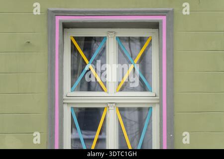 Double fenêtre blanche avec croix de ruban bleu et jaune, entourée de garnitures grises et roses sur le mur extérieur vert, détail de rue géométrique abstrait Banque D'Images