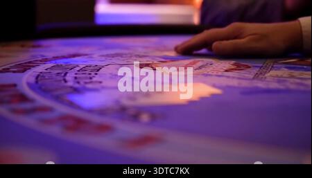 Main touchant une table de jeu dans un casino pendant la nuit avec des lumières colorées éclairant la scène et les joueurs à proximité placer des Paris Banque D'Images