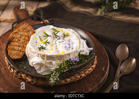 Le camembert fondu chaud est garni de brins de romarin frais et de fleurs comestibles. Cette photographie de cuisine rustique présente un éclairage naturel doux A. Banque D'Images