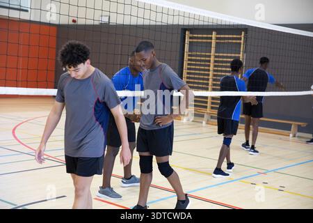 Coéquipiers de volleyball masculin adolescent pratiquant dans la salle de gym de l'école sous filet sur un court en bois avec genouillères Banque D'Images