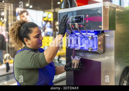 Femme dans des gants versant chocolat et vanille soft serve dans une tasse d'une machine à dessert glacé élégant derrière un comptoir de magasin, servant les clients dans un Banque D'Images