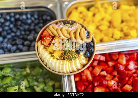 Bol à smoothie Acai avec des tranches vibrantes de banane, mangue, kiwi, fraises et bleuets, surmonté de tourbillons de beurre d'arachide, granola et pollen d'abeille pour Banque D'Images