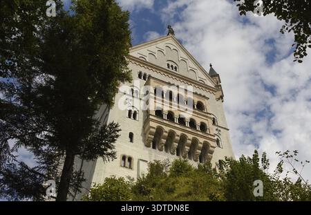 Château Neuschwanstein : une vue vers le haut d'une tour de château en pierre blanche avec des fenêtres cintrées entourées de grands arbres sous un ciel nuageux Banque D'Images