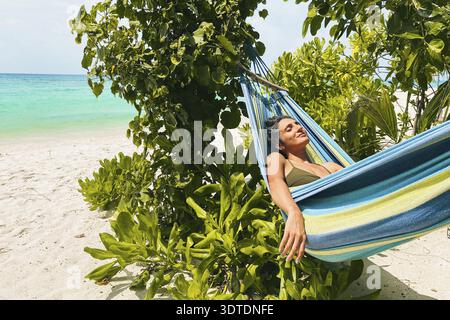 Femme brune hipster dans le hamac se détendre profiter du bain de soleil à l'extérieur dans la plage tropicale en plein air par l'océan en vacances aux Maldives. Blogger voyage infl Banque D'Images
