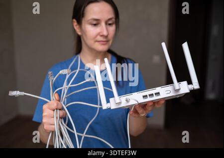 Jeune femme dans un t-shirt bleu tenant un routeur sans fil moderne et un tas de câbles Internet gris. Concept d'installation de réseau domestique, dépannage Banque D'Images