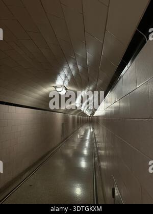 Long couloir de tunnel piétonnier éclairé avec murs carrelés et plafonniers créant de fortes lignes de perspective dans un passage souterrain. Banque D'Images
