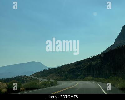 Autoroute sinueuse pittoresque à travers les montagnes désertiques arides du sud-ouest américain, avec des falaises rocheuses spectaculaires et des collines lointaines sous un ciel bleu clair. Banque D'Images
