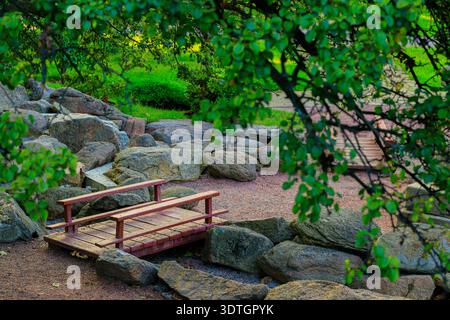 Un charmant pont en bois rustique avec des balustrades rouges enjambe un lit de ruisseau sec entouré de grandes pierres naturelles et d'un feuillage vert vibrant Banque D'Images