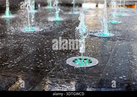 Des jets d'eau jaillissent des fontaines au sol, éclairés par des lumières vertes et bleues brillantes. La surface mouillée reflète les couleurs chatoyantes, créant un Banque D'Images