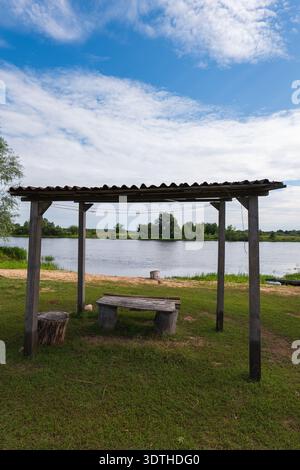 Un abri rustique avec des poteaux en bois, un toit ondulé, un banc, et des sièges de souche se dresse sur une rive herbeuse près d'une rivière calme bordée d'arbres sous un clou doux Banque D'Images