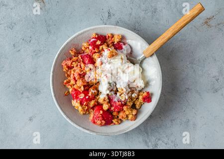Crumble de fraises avec crème glacée fondante, granola cuit au four, graines de citrouille et amandes dans un bol avec cuillère sur fond gris. Dessert aux fruits croustillants d'été Banque D'Images