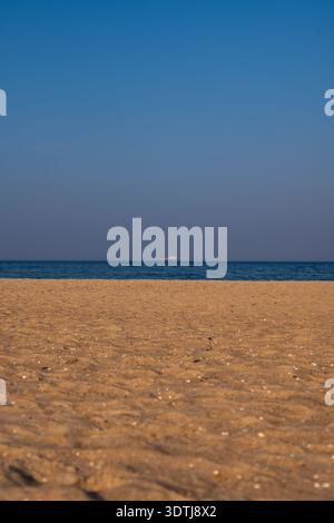 Une plage de sable calme rencontre une mer bleu profond calme sous un ciel clair, avec un seul navire à l'horizon. Des reflets lumineux chauds en fin d'après-midi sur les coquillages, minim Banque D'Images