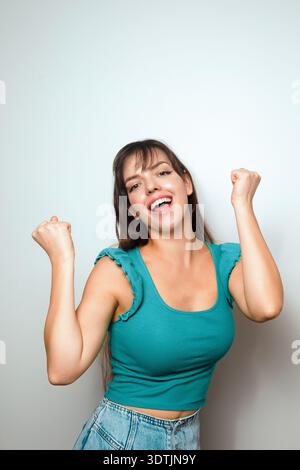 Une femme sourit largement en levant les poings pour célébrer. Elle se tient dans une pièce lumineuse aux murs Unis. La scène capte sa joie et son énergie. Banque D'Images