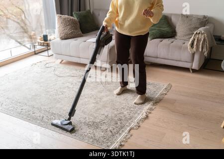 Une femme en vêtements décontractés est vue passer l'aspirateur sur un grand tapis beige dans un cadre confortable de salon. L'espace dispose d'un canapé confortable orné de va Banque D'Images