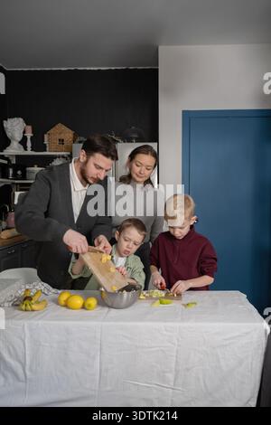 Une famille de quatre prépare une collation aux fruits saine dans leur cuisine. Les parents aident les enfants à hacher des bananes et des pommes sur une coupe en bois B. Banque D'Images