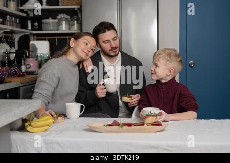 Une famille de trois prend son petit déjeuner dans une cuisine confortable. Les parents et l'enfant, tous souriants, s'assoient autour d'une table avec des tasses et des tranches de pain, en nourrissant Banque D'Images