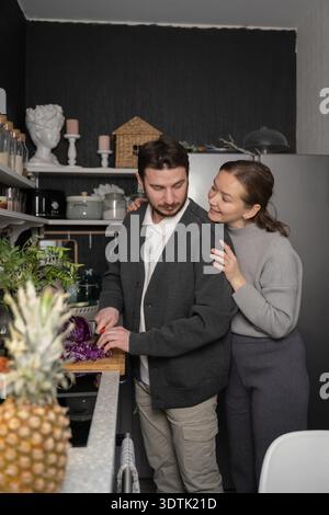 Un couple aime le temps de qualité à préparer un repas dans leur cuisine confortable. L'homme hache des légumes tandis que la femme l'embrasse affectueusement, highlightin Banque D'Images