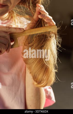 Gros plan vertical d'une femme peignant les longs cheveux blonds emmêlés avec un peigne en bois, montrant la rupture et les dommages. Conception des problèmes de soins capillaires pour dermatologue Banque D'Images