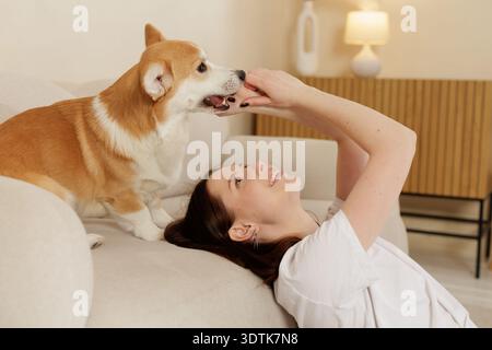 Jeune femme se relaxant sur un canapé tout en jouant avec son chien Corgi. Moment intérieur confortable montrant l'affection, la confiance et la joyeuse compagnie entre animal et Banque D'Images