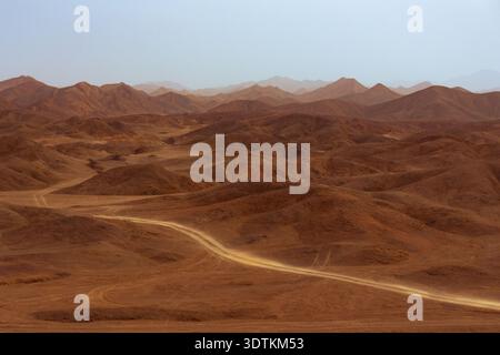 Vue panoramique sur un chemin de terre sinueux à travers un vaste paysage désertique aride avec des collines ondulantes et des montagnes lointaines sous un ciel poussiéreux. Banque D'Images