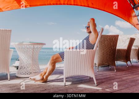 Vacances homme de détente de plage en maillot de bain vintage rayé assis sur une chaise en osier blanc à la terrasse du café de la mer sous la canopée rouge vif Banque D'Images