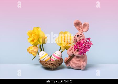 Lapin tricoté de Pâques rose avec fleur de jacinthe rose, nid d'oiseau avec des oeufs rayés roses et jaunes et fleurs printanières de jonquilles jaunes sur fond dégradé rose et bleu. Concept de Pâques Banque D'Images