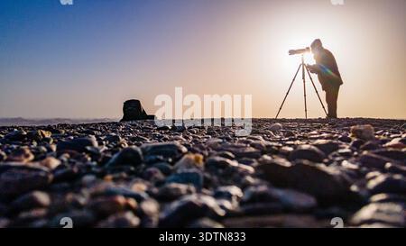 Silhouette d'un photographe utilisant un appareil photo monté sur trépied dans un paysage désertique au lever du soleil. Banque D'Images