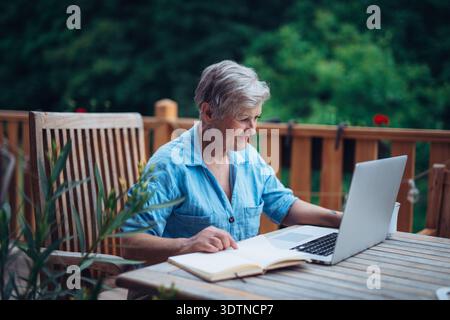 Femme mature assise sur le balcon avec ordinateur portable, travaillant à distance. Banque D'Images