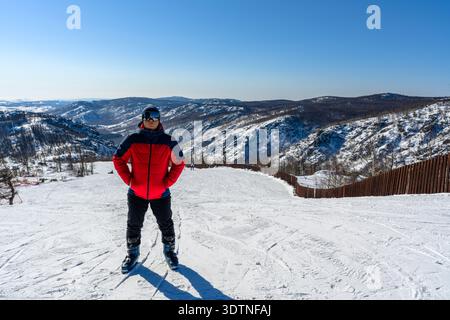 Un homme en costume de ski rouge, lunettes et bottes sur fond de piste de ski et de montagnes. Le concept de ski, sport, style de vie actif. Banque D'Images