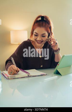 Une jeune femme joyeuse aux cheveux roux est assise à un bureau dans son bureau à domicile, parlant sur un smartphone tout en écrivant dans un planificateur en spirale, illustrant Banque D'Images