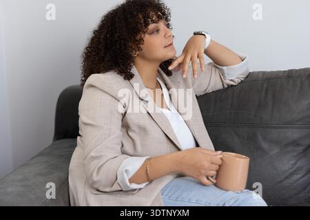 Femme afro-américaine assise sur un canapé gris tenant une tasse beige, portant un blazer et une smartwatch Banque D'Images