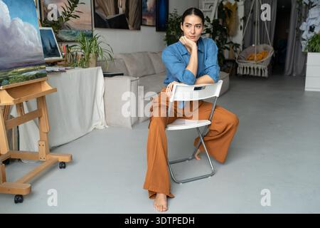 Artiste femme réfléchie dans le processus de pensée regardant à distance assis dans l'atelier de galerie loft entouré par l'affichage d'œuvres d'art. Méditant sur la femme peintre regardant la pensée de côté, obtenir de nouvelles idées. || autorisation du modèle Banque D'Images