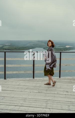 Femme rousse heureuse enveloppée dans un châle chaud se dresse sur la terrasse en bois surplombant le paysage pittoresque de la rivière avec des collines verdoyantes sous le ciel couvert pendant le vent Banque D'Images