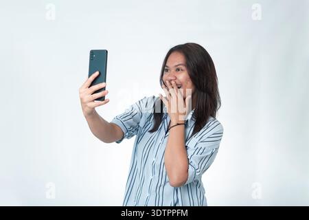 Femme indonésienne dans une chemise rayée bleue tenant son smartphone et couvrant sa bouche dans une expression joyeuse et surprise. En regardant l'écran du téléphone Banque D'Images