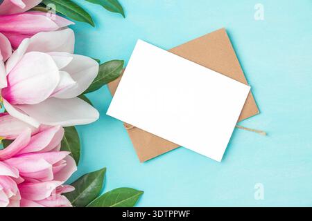 Fleurs de magnolia rose et feuilles vertes avec carte de voeux vierge et enveloppe sur fond bleu pastel. Maquette avec espace de copie. Pose à plat. Mariage Banque D'Images