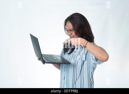 Femme indonésienne portant des lunettes et une chemise rayée regardant un ordinateur portable avec une expression confuse et ennuyée tout en pointant vers l'écran. Isolé Banque D'Images