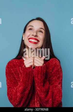 La joie rayonne d'une jeune femme portant un pull rouge confortable. Elle sourit largement, montrant son excitation et son bonheur, capturés contre un b bleu vif Banque D'Images