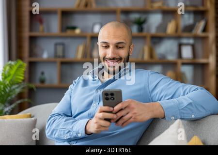 Jeune homme se détendant sur un canapé, regardant son smartphone, se connectant avec ses amis et sa famille, et profitant de la communication numérique et du divertissement dans un environnement domestique moderne Banque D'Images