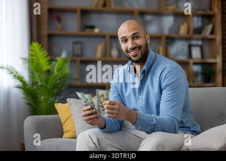 Jeune homme joyeux souriant, tenant des piles de billets de banque en dollars, gérant avec succès son budget et comptant l'argent tout en étant assis sur un canapé confortable dans un salon moderne Banque D'Images