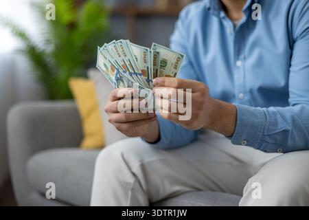 Homme dans des vêtements décontractés assis sur un canapé, tenant un ventilateur de billets de cent dollars dans ses mains, représentant des concepts de finance, revenu, richesse, budget, profit, et le succès Banque D'Images