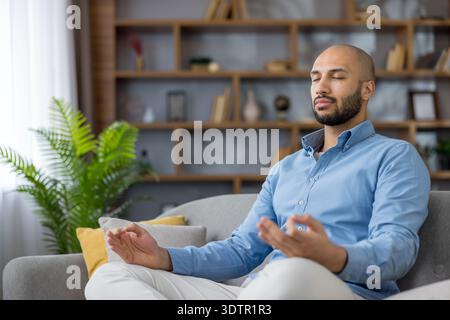 Jeune homme assis confortablement sur un canapé à la maison, pratiquant la méditation de pleine conscience avec les yeux fermés et les mains dans le mudra, trouvant le calme et gérant le stress pour le bien-être mental Banque D'Images