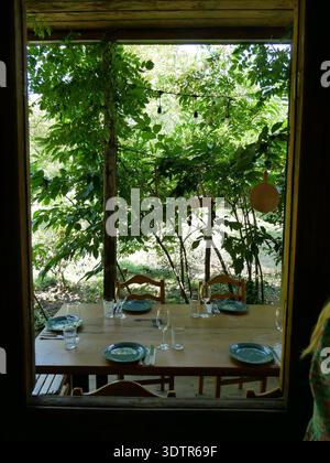 Une table dressée pour le déjeuner sous une pergola feuillue, vue à travers la fenêtre d'une maison traditionnelle, Imereti, Géorgie, juillet 2023. Banque D'Images