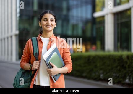 Heureux étudiant indien portant un sac à dos et des cahiers, marchant en toute confiance sur un campus universitaire, représentant l'éducation, la réussite et les aspirations professionnelles futures Banque D'Images
