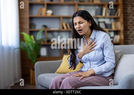 Jeune femme ressentant une douleur thoracique aiguë et des difficultés respiratoires alors qu'elle était assise sur un canapé dans son salon, indiquant une crise cardiaque ou une crise d'anxiété potentielle Banque D'Images