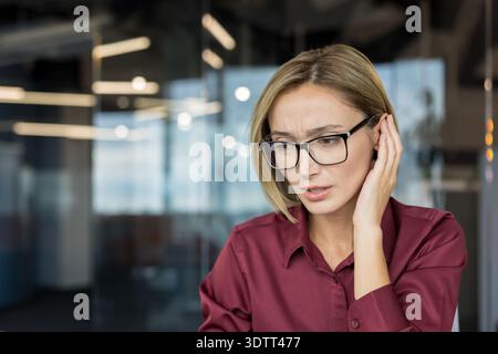 Femme d'affaires adulte dans des lunettes caressant l'oreille, luttant pour entendre ou comprendre une conversation dans un bureau moderne, représentant des concepts d'audition difficul Banque D'Images