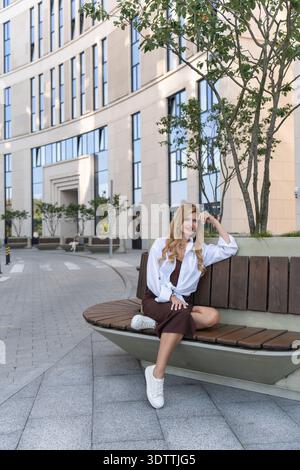 Portrait d'une femme d'affaires se reposant à l'extérieur pendant sa journée de travail, entourée d'une architecture propre et de verdure. Banque D'Images