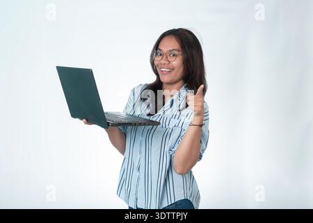 Joyeuse femme indonésienne portant des lunettes et une chemise rayée bleue, tenant un ordinateur portable dans une main et pointant son autre index vers le spectateur Banque D'Images