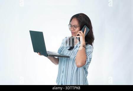 Femme indonésienne portant des lunettes et une chemise rayée bleue, semblant stressée et concentrée tout en tenant un ordinateur portable et en parlant sur son smartphone à la sa Banque D'Images